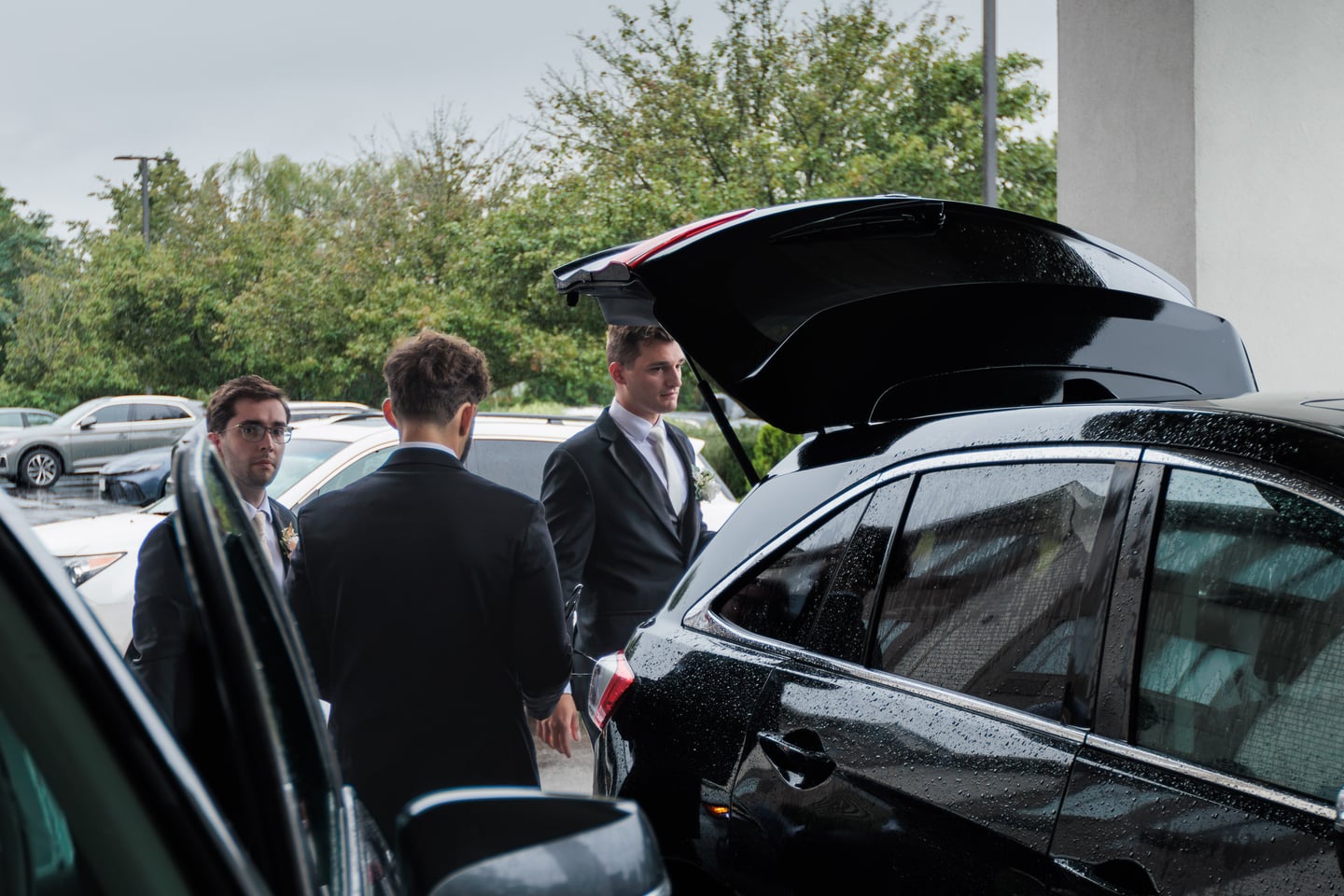 Men in suits stand beside an open car trunk before departing.