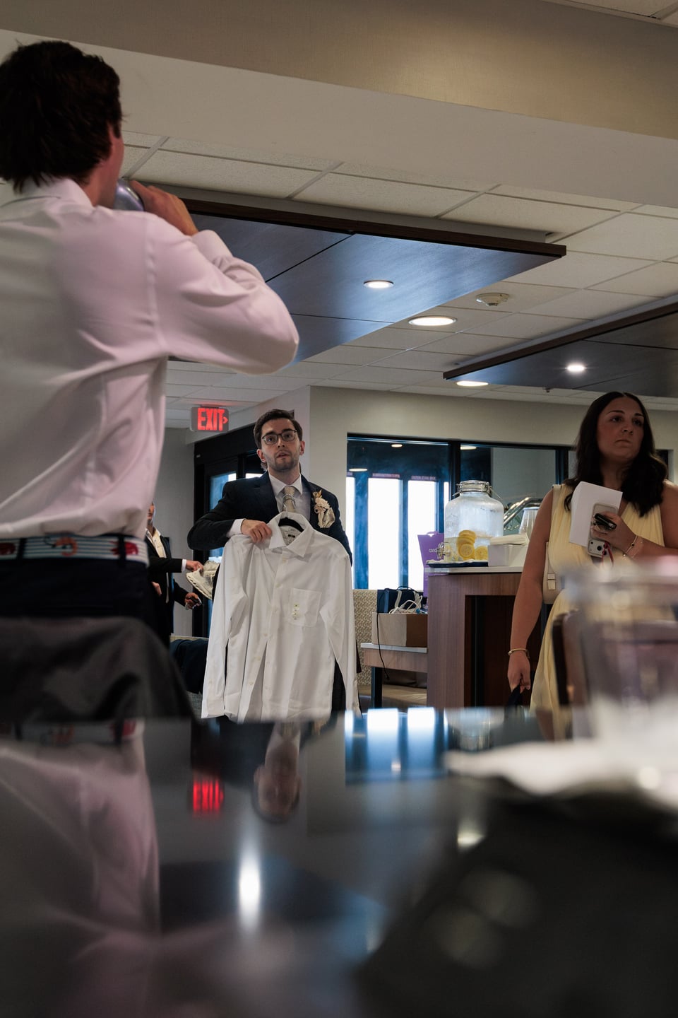 A guest stands near a mirror as the wedding party gets ready in a hotel room.