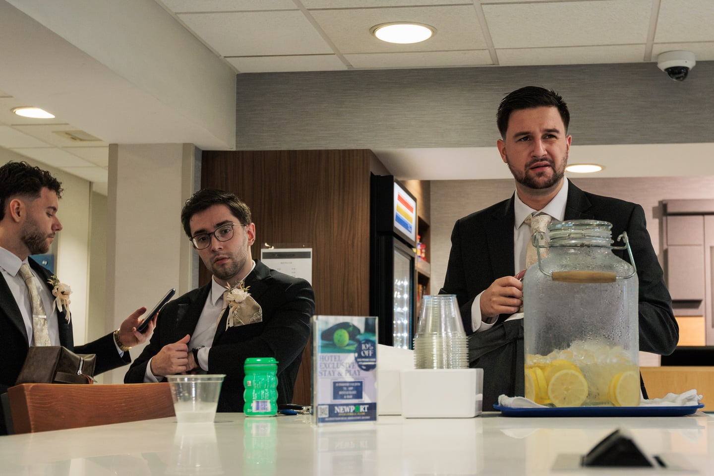 A man in a dark suit holds a drink while others gather nearby.