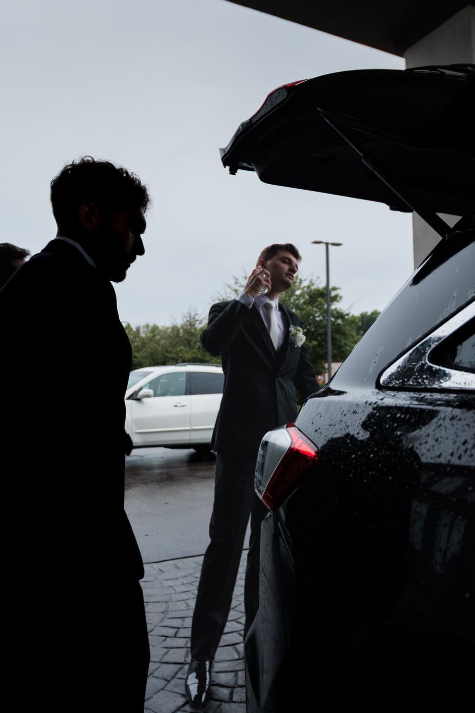 Silhouetted wedding guests stand beside a car with its trunk open.