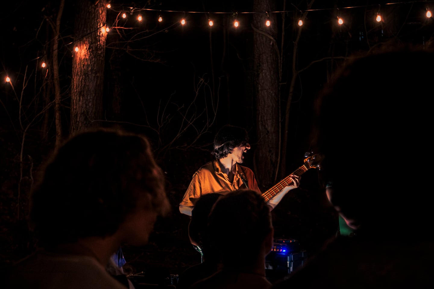 A bassist in an orange shirt plays beneath string lights beyond dark audience silhouettes.