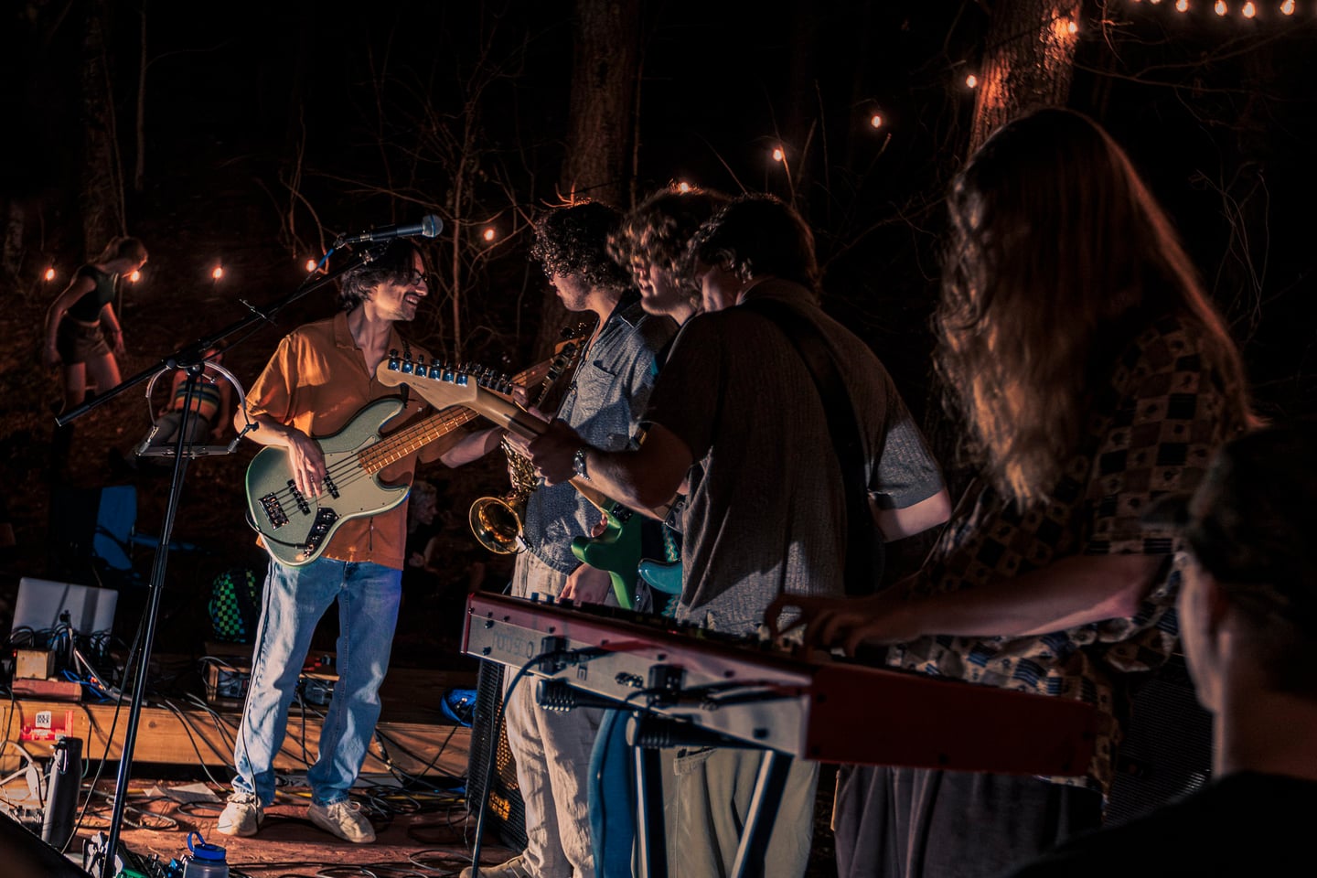 Band members smile toward one another while playing bass, guitars, and keyboard.