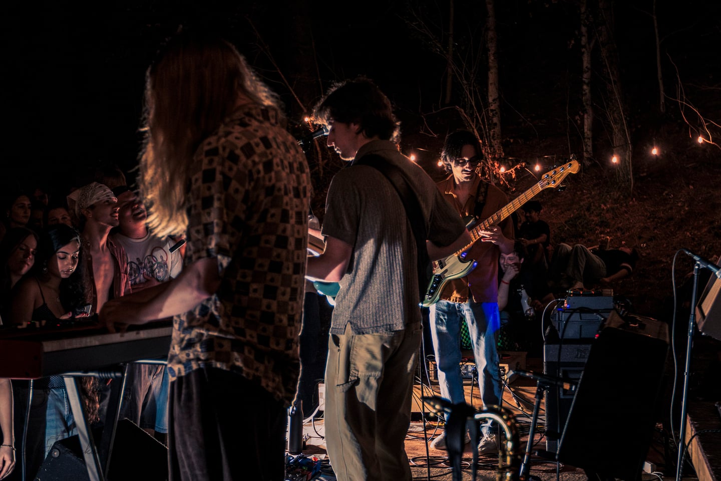 A bassist and guitarist perform among musicians and audience members under string lights.