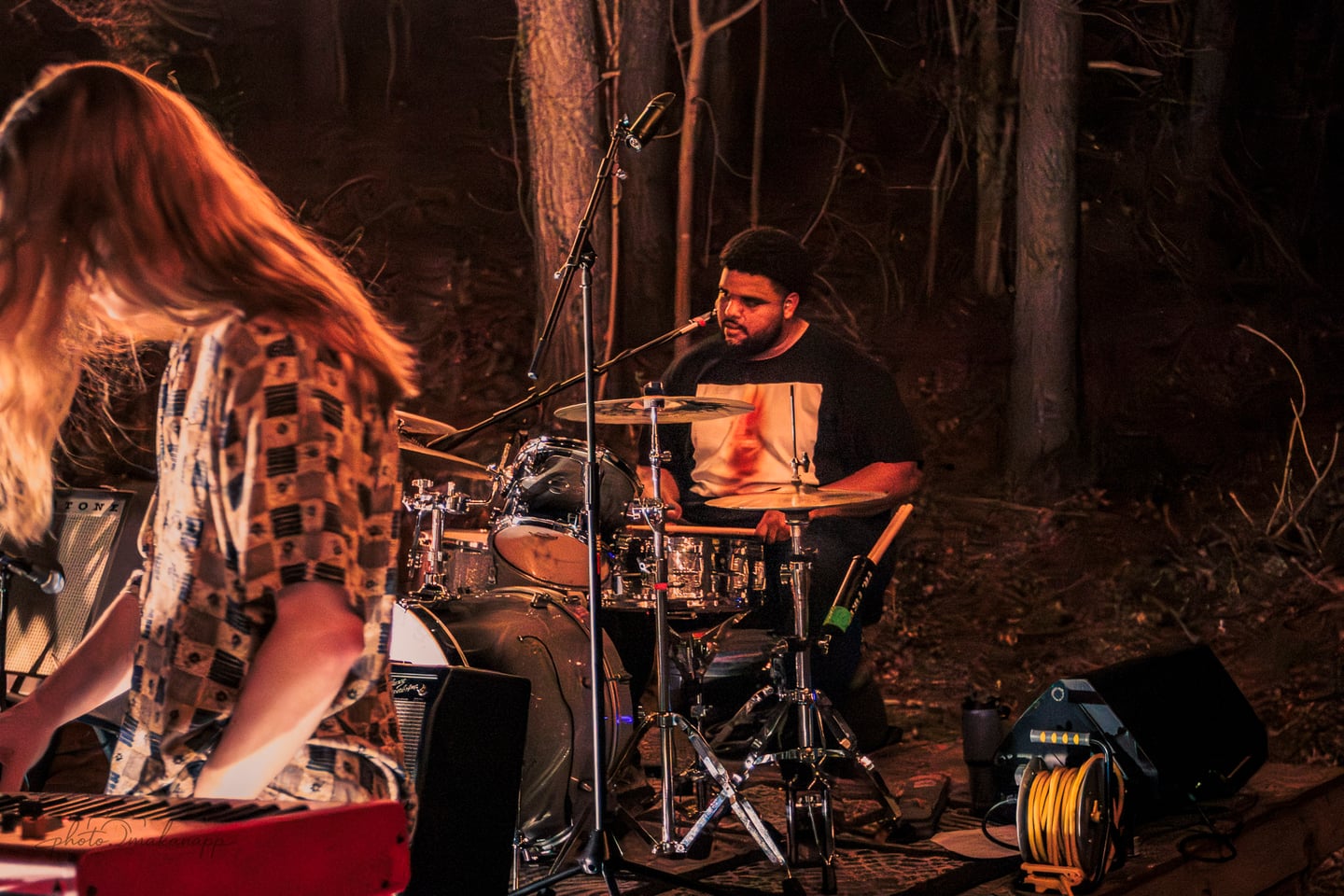 The drummer plays behind the keyboardist in warm stage light among the trees.