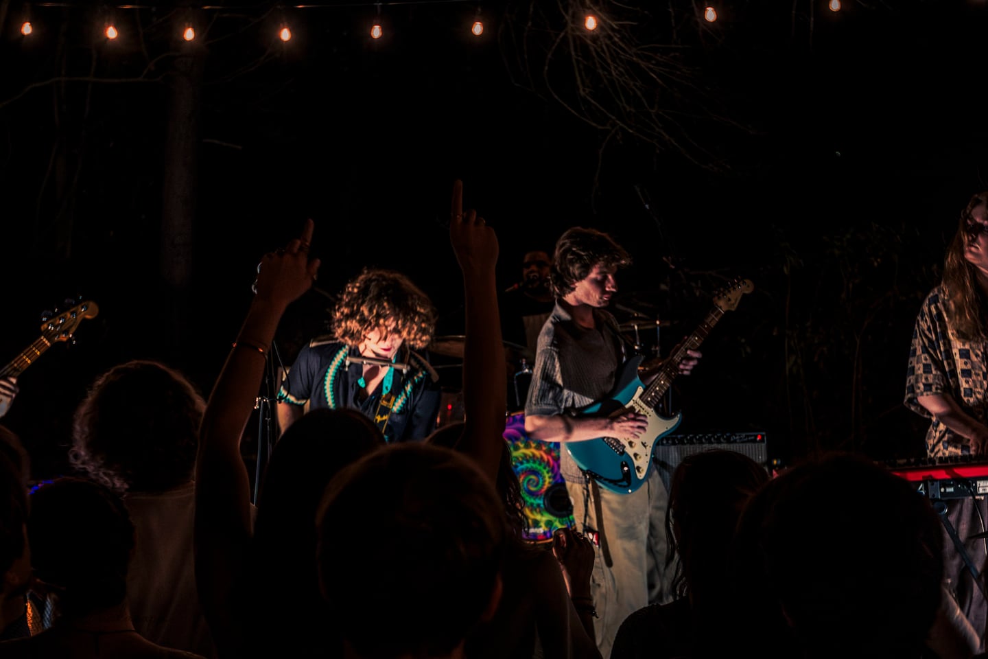 Two guitarists and a keyboardist perform behind audience members with their arms raised.