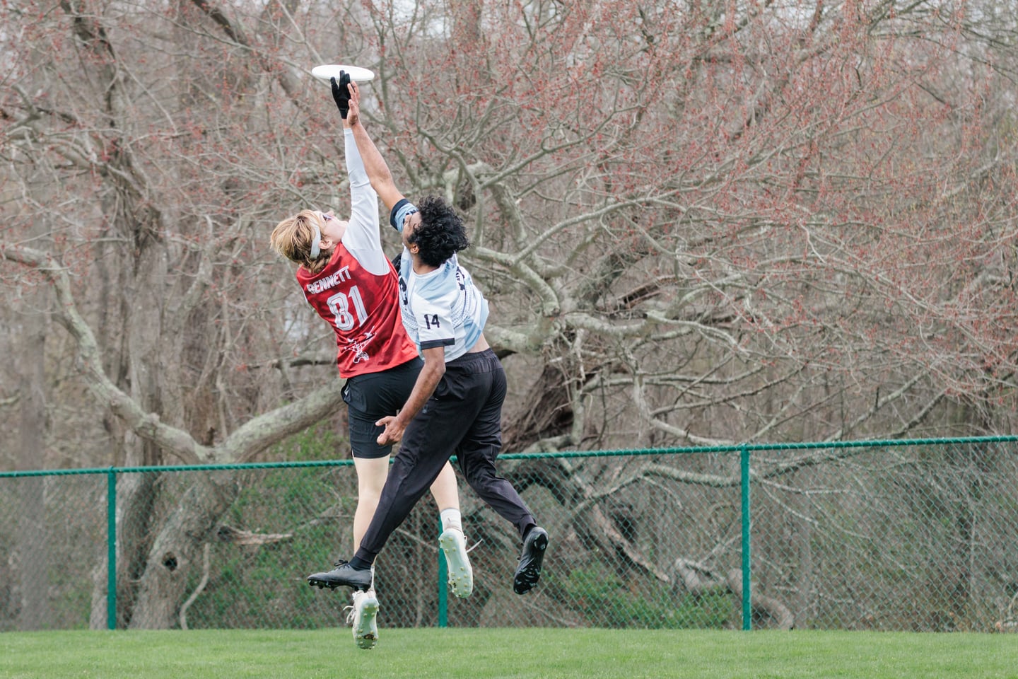 Red-clad player and dark-clad opponent leap together for an overhead disc.