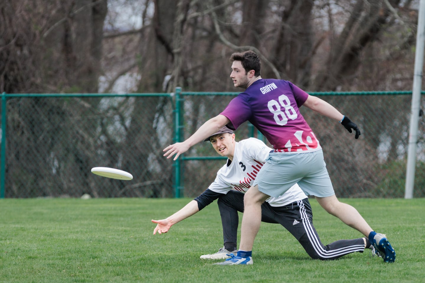 White-clad player lunges low toward the disc as a purple-clad defender closes.