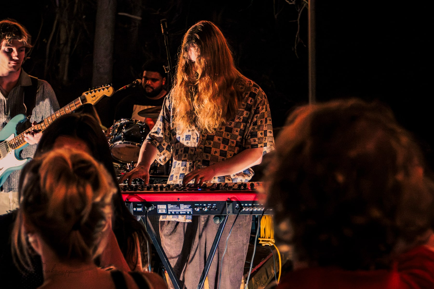 A keyboard player performs behind a red keyboard while audience members frame the foreground.