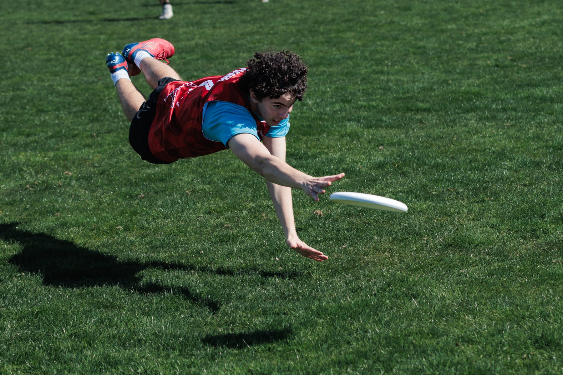 A red-shirted player dives horizontally toward a low-flying disc over the grass.